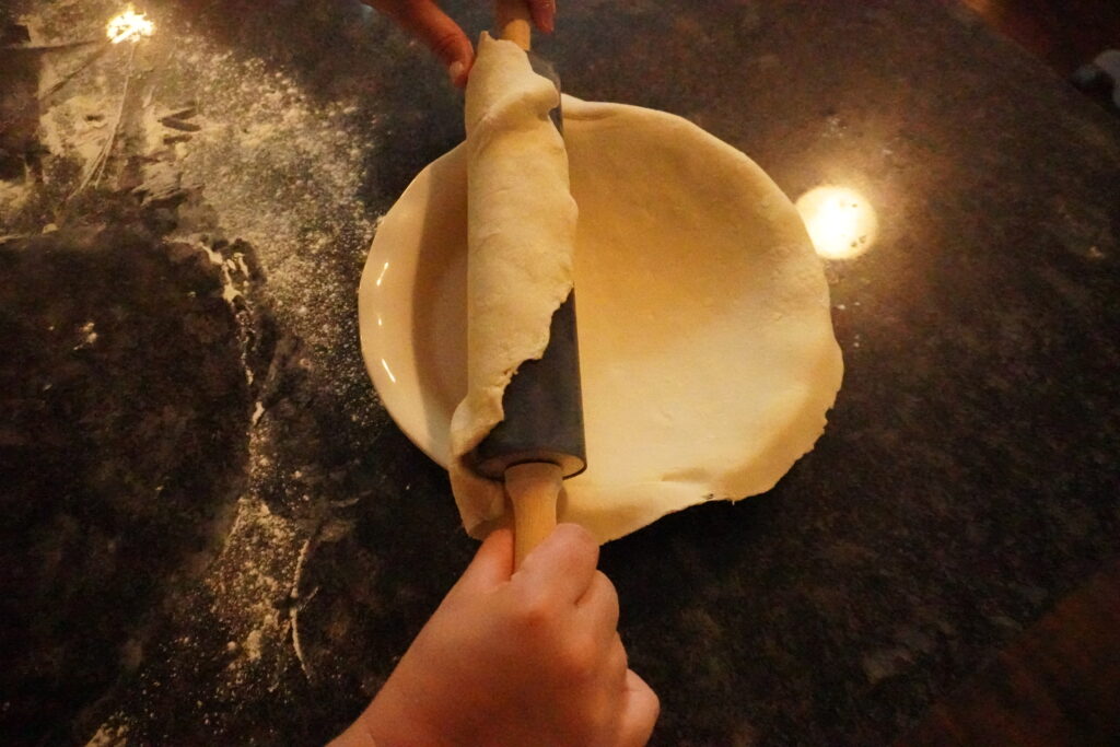 pie dough being unfurled with a rolling pin onto a white pie plate